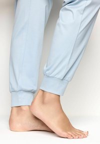 Pies descalzos con pantalones jogger de color azul claro arremangados en los tobillos, mostrando las uñas de los pies pintadas de rosa pálido sobre un fondo blanco.