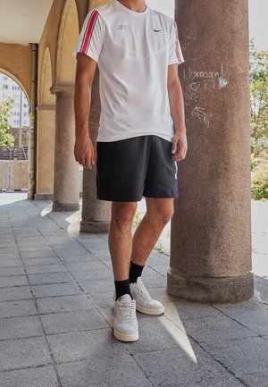 Un homme se tient à l'extérieur, portant un t-shirt blanc Nike avec des rayures rouges, un short noir, des chaussettes noires et des baskets blanches sur une allée carrelée avec des colonnes.