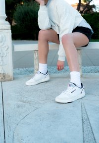 Vita lädersneakers med marinblå accenter och röd logotyp, kombinerade med vita strukturerade crew-strumpor, placerade på en mönstrad betongyta.