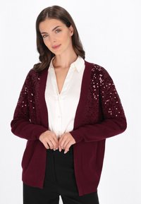 Bordo kardigan z ozdobnymi cekinami na ramionach, z długimi rękawami, otwartym przodem oraz prążkowanymi mankietami i dołem. W zestawie z białą koszulą z guzikami.