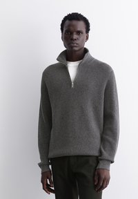 Grau gestreifter Strickpullover mit Reißverschlusskragen, langen Ärmeln und tailliertem Saum. Getragen über einem weißen Hemd mit strukturiertem Finish.