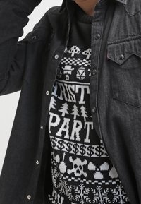 Camicia in denim nera con bottoni indossata sopra un maglione nero e bianco con motivi a tema natalizio, tra cui alberi e un design di teschio.