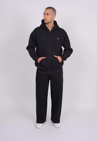 Felpa nera con zip e tasche a marsupio, abbinata a pantaloni sportivi a gamba larga coordinati. Entrambi i capi presentano un dettaglio di logo minimalista.