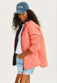 Prošívaná korálová bunda s černými akcenty a předním zipem, spárovaná se světle modrými džíny. Modrá kšiltovka s nápisem "LOS ANGELES" v bílé barvě.