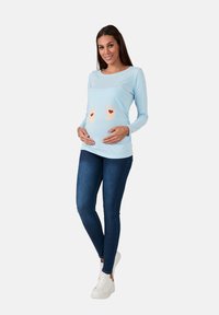 Helles blaues Langarmoberteil mit einem Bauchgrafik, die gelbe Babyfüße und rote Herzen zeigt, kombiniert mit dunkelblauen enganliegenden Jeans und weißen Turnschuhen.