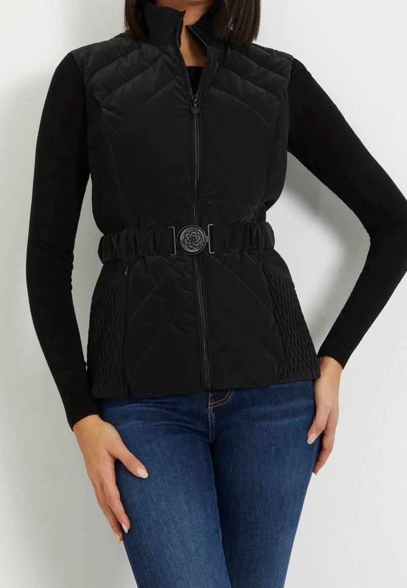 Femme portant un gilet matelassé sans manches noir avec une ceinture à boucle florale par-dessus un haut noir à manches longues et un jean bleu.
