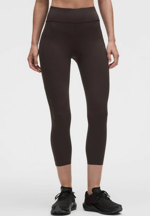 Frau trägt schwarze kurze Leggings und schwarze Sportschuhe und steht vor einem schlichten Hintergrund.