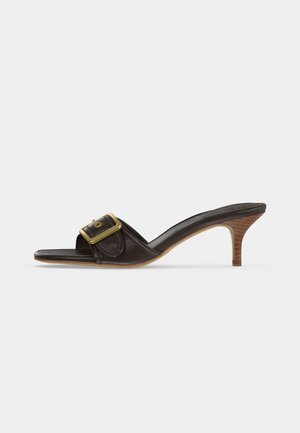 Mule noire à bout ouvert avec un petit talon marron empilé et une large boucle dorée sur la bride au-dessus du pied.