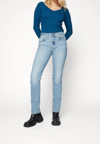 Jeans de mezclilla de tono azul claro, de cintura alta y corte ajustado, combinados con una blusa de manga larga ceñida en color teal y botas de tobillo negras con tacón.