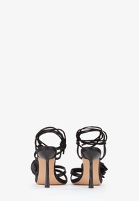 Sandali neri con tacco alto caratterizzati da un design a fascia con lacci intrecciati alla caviglia, una texture in satin liscio e un tacco a stiletto con una suola beige.