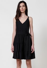 Schwarzes ärmelloses Kleid mit V-Ausschnitt, gestuftem Rüschensaum und gebundenem Taillendetail, aus strukturiertem Stoff mit subtilen Mustern.