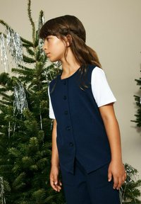 Temno modra telovnik s pokončnimi črtami, zapenjan z gumbi, oblečen čez belo majico, stoji ob božičnem drevesu z okraski iz lameta.