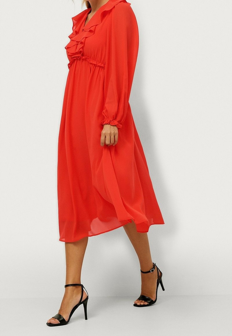 Robe en chiffon rouge avec un col à volants, des manches longues et une taille cintrée, assortie à des sandales noires à talons hauts et à brides.