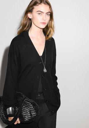 Black cardigan with a v-neck, buttoned front, and long sleeves. Paired with a black woven bag and layered silver necklace.