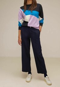 Pull coloré en violet, bleu et noir avec des motifs géométriques, associé à un pantalon large marine et des sneakers blanches.