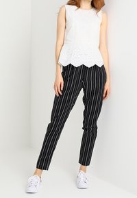 Top bianco senza maniche con dettagli a perforazione e orlo scallop, abbinato a pantaloni tapered a righe nere e sneakers bianche.