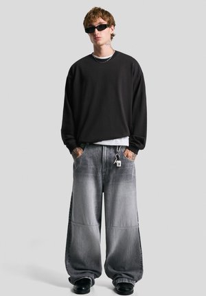 Junger Mann in einem schwarzen übergroßen Sweatshirt, weiten grauen Jeanshosen, schwarzen Schuhen, Sonnenbrille und Tätowierungen an Händen und Nacken, der steht.