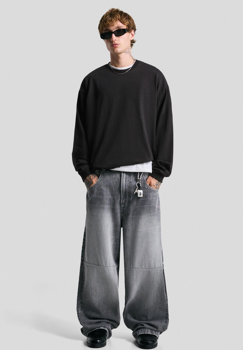 Jeune homme portant un sweatshirt noir surdimensionné, un pantalon en denim gris large, des chaussures noires, des lunettes de soleil, et des tatouages sur les mains et le cou, debout.