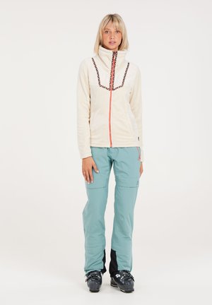 Veste en polaire crème avec un col décoratif, associée à un pantalon cargo bleu clair présentant plusieurs poches et des accents noirs.