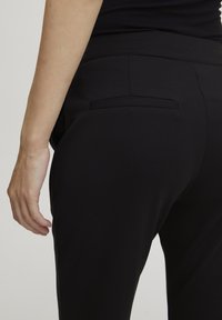 Zwarte rekbare stoffen broek met een naadloze tailleband, gestroomlijnd ontwerp en één achterzak, gezien vanuit de zijkant.