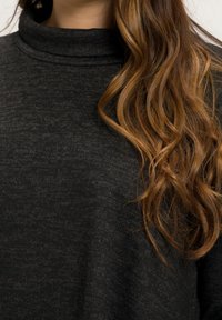 Dark gray, textured turtleneck sweater with a high collar. Soft fabric, relaxed fit. Model's wavy, long hair partially visible.