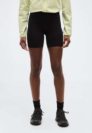 Άτομο που φοράει μαύρα αθλητικά σορτς μέχρι το μέσο του μηρού, μαύρες κάλτσες, μαύρα αθλητικά παπούτσια και ένα ανοιχτό κίτρινο μπουφάν με φερμουάρ και μακριά μανίκια, στέκεται μπροστά σε λευκό φόντο.