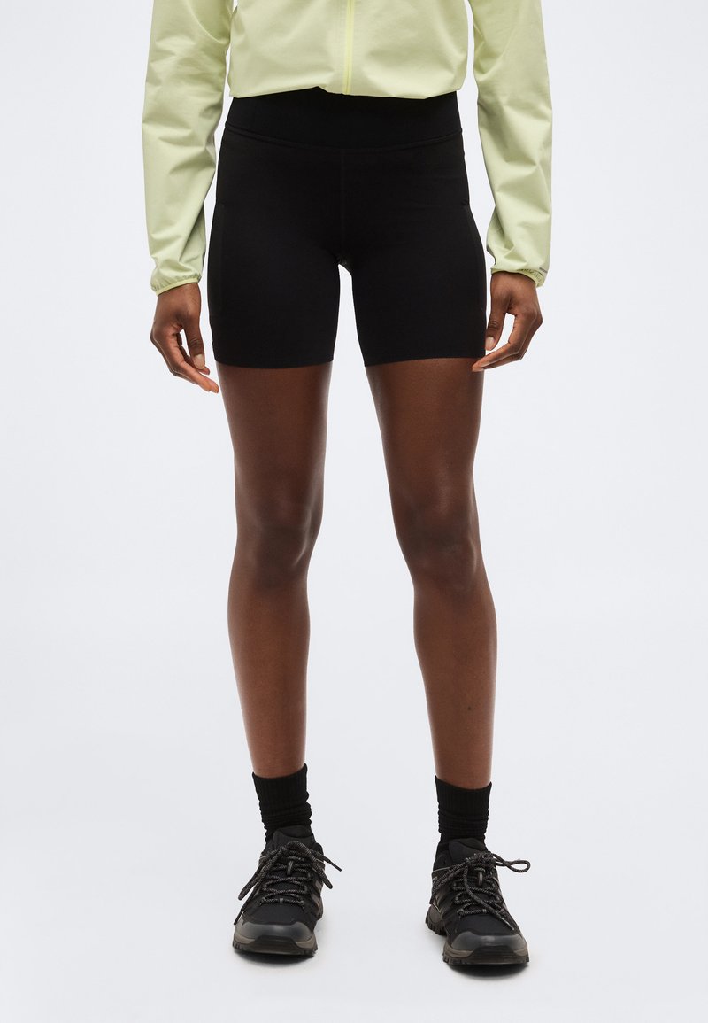 Άτομο που φοράει μαύρα αθλητικά σορτς μέχρι το μέσο του μηρού, μαύρες κάλτσες, μαύρα αθλητικά παπούτσια και ένα ανοιχτό κίτρινο μπουφάν με φερμουάρ και μακριά μανίκια, στέκεται μπροστά σε λευκό φόντο.