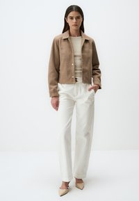 Veste en laine brune courte avec un col, fermeture à boutons et deux poches latérales. Associée à un pantalon large blanc et des chaussures beige à bout pointu.