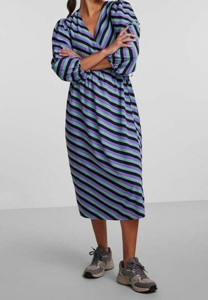 Nainen pukeutuneena midi-pituiseen mekkoon, jossa on diagonaalisia mustia, violetteja ja vihreitä raitoja, pikeerityt pitkät hihat, yhdistettynä harmaisiin lenkkareihin.