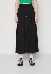 Musta maksihame, joka on valmistettu tekstuurikankaasta, etuosassa napitus ja leveä helma. Yhdistetty kiiltäviin hopeisiin solkikenkiin ja valkoisiin sukkiin.