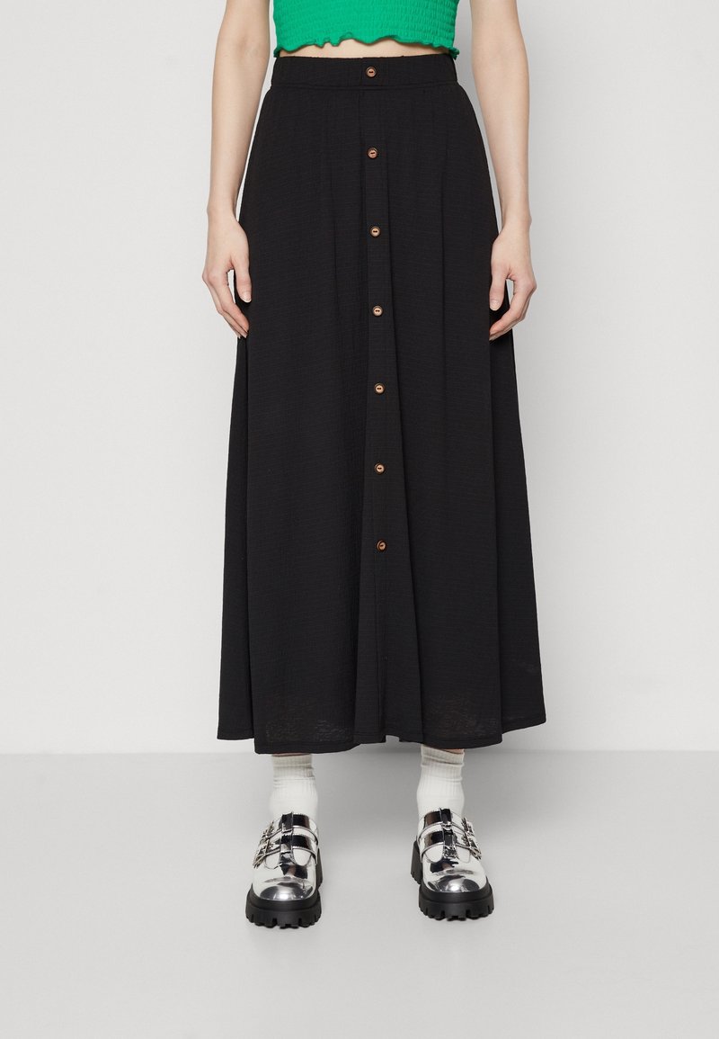 Musta maksihame, joka on valmistettu tekstuurikankaasta, etuosassa napitus ja leveä helma. Yhdistetty kiiltäviin hopeisiin solkikenkiin ja valkoisiin sukkiin.