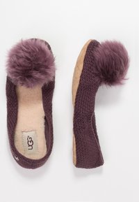 Fioletowe, dziane kapcie z miękkim, puszystym pomponem, z kremowym futrzanym wykończeniem i beżową zamszową podeszwą. Logo UGG widoczne w środku.