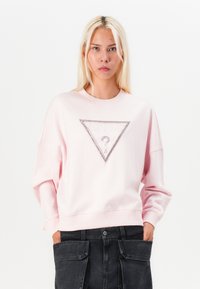 Light pink sweatshirt with a triangular "GUESS" logo. Features rhinestone accents and dropped shoulders, paired with dark cargo pants.