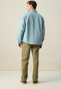 Chemise boutonnée bleu clair à manches longues, associée à un pantalon vert olive et des sandales marron. La chemise présente un empiècement au dos et des fentes sur les côtés.