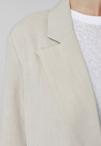 Gros plan d'une personne portant un blazer en lin beige clair sur un t-shirt blanc texturé à col rond, montrant les détails du col et de l'épaule.