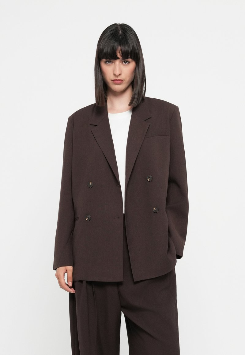 Brązowy dwurzędowy żakiet z klapami i dwoma przednimi kieszeniami. Gładki materiał z luźnym krojem, zestawiony z białym t-shirtem pod spodem.