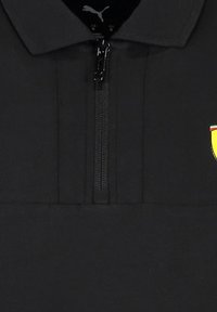 Czarna koszulka polo z ćwierć zamkiem. Zawiera małe, haftowane logo tarczy w odcieniach żółtego i czerwonego. Gładka tekstura, wytrzymały materiał.