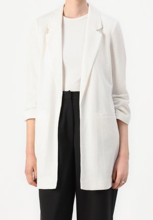 Personne portant un blazer blanc texturé sur un haut côtelé blanc, associé à un pantalon noir ample taille haute, debout devant un fond blanc.