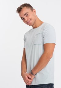 T-shirt grigio chiaro a maniche corte con taschino sul petto, scollatura rotonda e texture in cotone morbido. Indossato con jeans blu navy, mani unite.