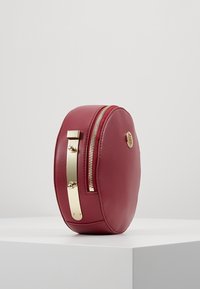 Borsa rotonda in pelle bordeaux con zip dorata e dettagli in oro. Presenta una chiusura con un moschettone piatto dorato al lato. Texture liscia.