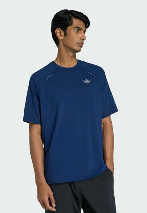 Mann trägt ein blaues Adidas-Kurzarmshirt mit Logo auf der Brust, schaut zur Seite, Hände entspannt, steht vor einem schlichten Hintergrund.