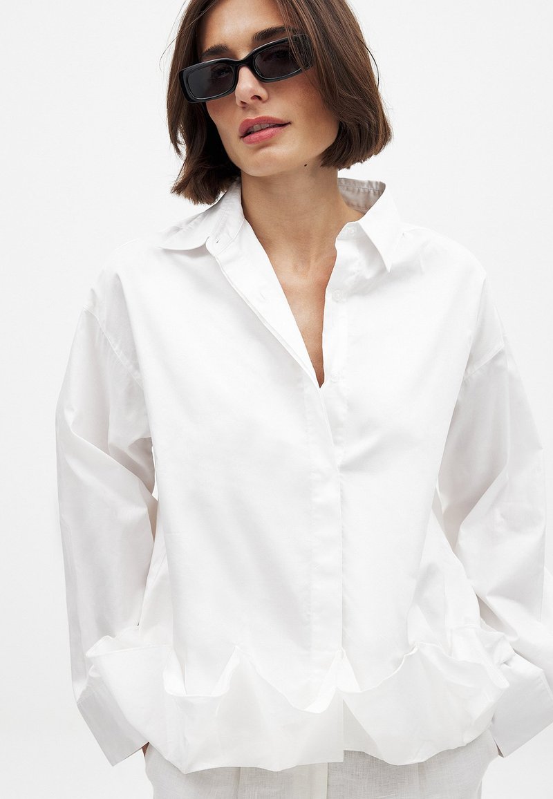 White cotton shirt with a ruffled hem, collared neckline, and long sleeves, featuring a smooth texture and relaxed fit.