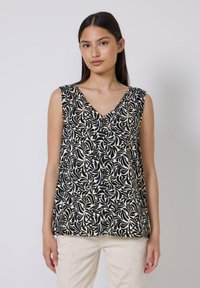 Vrouw in een mouwloze beige top met zwart bladerenpatroon en lichte broek, staand tegen een effen lichte achtergrond.
