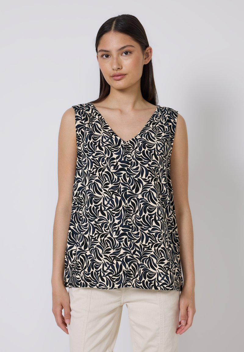 Vrouw in een mouwloze beige top met zwart bladerenpatroon en lichte broek, staand tegen een effen lichte achtergrond.