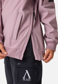 Jasnoróżowa kurtka na zamek błyskawiczny z prążkowanymi mankietami i dołem, wyposażona w boczne kieszenie oraz kontrastowe czarne logo na spodniach pod spodem.