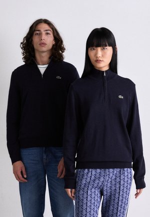 Due modelli stanno fianco a fianco indossando felpe navy a zip con logo di coccodrillo verde; uno indossa jeans blu, l'altro pantaloni a fantasia.