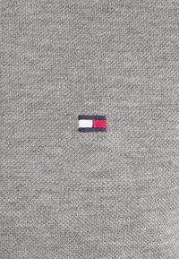 Grå stickad sweatshirt med en liten broderad logotyp i rött, vitt och marinblått, placerad på vänster sida. Mjuk textur.