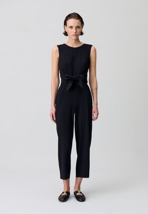 Combinaison sans manches noire avec une taille cintrée et un détail en nœud, présentant des jambes fuselées et une texture lisse. Portée avec des chaussures plates noires.