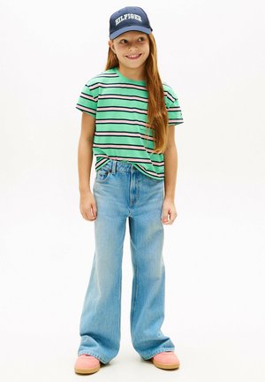 Giovane con capelli lunghi che indossa una t-shirt verde a righe, jeans larghi blu, scarpe rosa e un cappellino blu scuro con la scritta "HILFIGER".