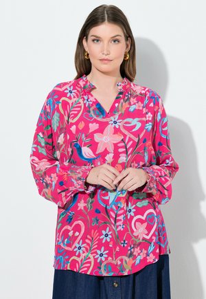 Kobieta w jasnoróżowej bluzce w kwiaty z niebiesko-zielonymi liśćmi, ptakami i wzorami serc, uzupełniona złotymi kolczykami kołami.